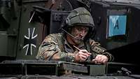 Ein Soldat mit Helm und Headset steht in der Dachluke eines Panzers und schreibt etwas auf.