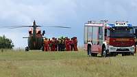 Ein Feuerwehrfahrzeug und ein Helikopter stehen auf einer Wiese, dazwischen Personen in Uniformen