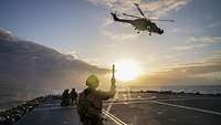 Soldaten auf dem Flugdeck einer Fregatte bei tief stehender Sonne. Über ihnen fliegt ein Hubschrauber.