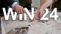 A wargame map is lying on the table. In front of the ‘Win 24’ lettering, two fingers are pointing towards playing pieces.