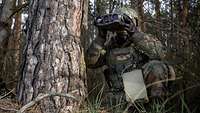 Ein Soldat hockt neben einem Baum und schaut mit einem Fernglas in die Ferne