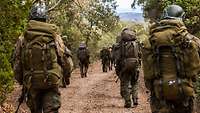 Soldaten mit großen Rucksäcken marschieren in Reihe auf einem sandigen Waldweg