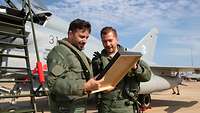 Two pilots look at a picture frame while a fighter jet is in the background.