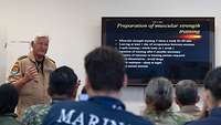 Ein Soldat steht neben einem Bildschirm mit einer Präsentation und spricht zu sitzenden Soldaten