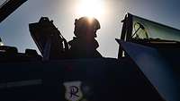 A pilot sits in the cockpit of an open fighter jet while the sun shines in the sky.