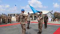 Ein Soldat salutiert vor einem anderen Soldaten mit UN-Flagge. Im Hintergrund weitere Soldaten.
