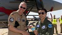 Two soldiers in flight suits stand in front of a fighter jet and hold a patch.