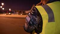 At an airport at night, the viewer looks over the shoulder of a soldier wearing a high-visibility vest.