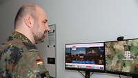 Ein Mann in Flecktarn steht am Schreibtisch und blickt auf zwei Monitore