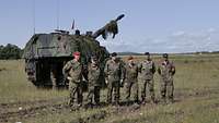 Fünf Soldaten und eine Soldatin stehen nebeneinander auf einem Feld vor einer Panzerhaubitze