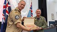 A German soldier and an Australian soldier in flight suits hold a wooden box in front of a stage.