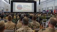 Soldiers wearing different uniforms sit in a room and look at a screen.