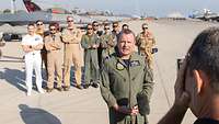 A pilot gives a press statement while other soldiers and fighter jets stand in the background.