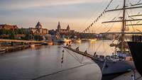 Mehrere Segelschiff liegen in einem breiten Fluss vor einer historischen Altstadt