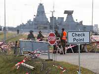 Mehrere Soldaten stehen hinter Absperrungen. Vor ihnen steht ein Schild, hinter ihnen eine Fregatte.