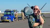 Ein Soldat in Pilotenkombi steht auf dem Rollfeld eines Flugplatzes vor einer großen grauen Drohne