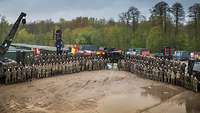 Mehrere Soldatinnen und Soldaten stehen angetreten auf einem Platz. Im Hintergrund stehen mehrere Container und Fahrzeuge. 