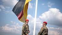 Zwei Soldaten stehen sich gegenüber vor einem Flaggenmast. Dabei wird die Flagge eingeholt.