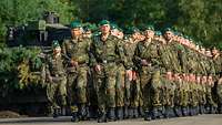 Soldaten marschieren in einer Antreteformation in drei Reihen, im Hintergrund ein Panzer