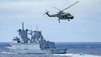  A gray warship at sea, with a gray helicopter in front of it.