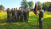 Eine Gruppe Soldatinnen und Soldaten stehen auf einer Wiese. Einer von ihnen trägt eine Fahne. 