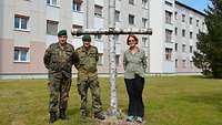 Zwei Soldaten und eine Pfarrerin stehen an einerm Birkenkreuz in vor einem Kasernengebäude.