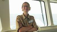 Eine Frau mit Brille in sandfarbener Uniform steht vor einem Schifffenster.