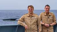 Zwei Männer in sandfarbener Uniform stehen neben einer Antenne auf einem Schiff.