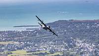 Ein Flugzeug fliegt über einer Küstenlandschaft, das Meer im Hintergrund.