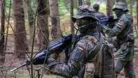 Drei Soldaten laufen mit Waffen im Anschlag nebeneinander durch den Wald.