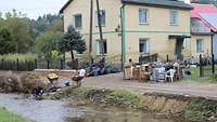 Ein Einfamilienhaus an einem schmalen Fluss mit deutlicher Flutmarke. Inventar steht am Straßenrand.