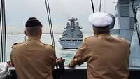 Zwei Männer in Uniform schauen einem grauen großen Schiff hinterher.