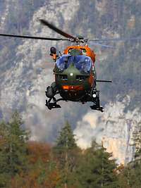 Ein Hubschrauber der Bergwacht im Flug über Bäume. Im Hintergrund ist eine Bergwand zu sehen.