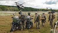 Auf einer Wiese stehen zwei Motorräder und Soldaten, im Hintergrund fliegen Hubschrauber.