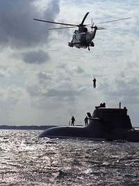 Ein Hubschrauber mit einer Person an der Winde über einem aufgetauchten U-Boot der Klasse U212.