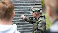 Ein Soldat in Tarnuniform deutet auf eine Stehle mit Namensbändern