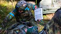 Ein Soldat, welcher mit einem roten Kreuz markiert ist, versorgt einen anderen Soldaten