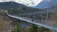 Eine Hängeseilbrücke, auf der zwei Menschen stehen, überbrückt ein Tal.