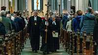 Geistliche gehen aus der Kirche, angeführt von einem Pfarrer, der ein Kreuz vor sich trägt.