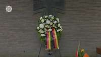 Ein Blumenkranz mit einer Deutschland-Kranzschleife vor einer Mauer auf der das Eiserne Kreuz ist.