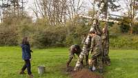 Fünf Personen in Uniform versenken ein Holzkreuz in einem Erdloch. Eine Frau steht vor ihnen.