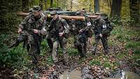 Mehrere Soldaten marschieren mit Gepäck und einer großen Kiste durch den Wald an Pfützen vorbei.