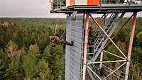 Ein hoher Turm steht in einem Wald und ein Soldat seilt sich mit Ausrüstung von dem hohen Turm ab.