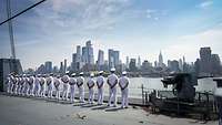 Soldaten in weißer Uniform aufgereiht auf einem grauen Schiff mit Blick auf New York.