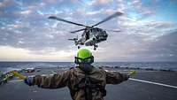 Eine Person mit Helm gibt Signale an einen Hubschrauber zur Landung auf einem Schiffsflugdeck.