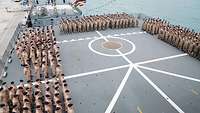 Viele Marinesoldaten in brauner Uniform sind auf dem Flugdeck eines Schiffes angetreten.