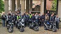 21 Menschen mit Motorradbekleidung stehen hinter vier Motorrädern.