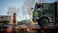 An einem Güterbahnhof steht ein Zug. Auf dem Waggon steht ein Soldat vor einem Lastwagen. 