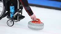 Ein Sportler im Rollstuhl sitzend stösst einen Curlingstein an