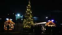 Ein erleuchteter Weihnachtsbaum steht zwischen zwei erleuchteten Buden auf dem Weihnachtsmarkt.